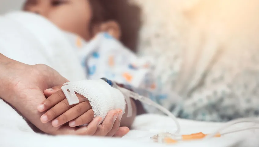 A small child lying in a hospital bed. An adult is holding the child's hand, which has an IV drip wrapped in gauze.
